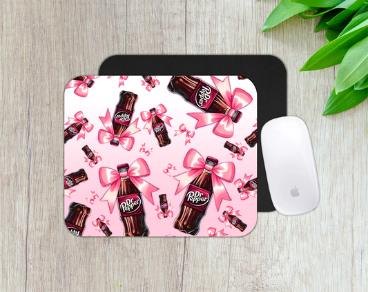 Mouse pad with Dr Pepper bottles and pink bows on a pink background, placed on a wooden surface.